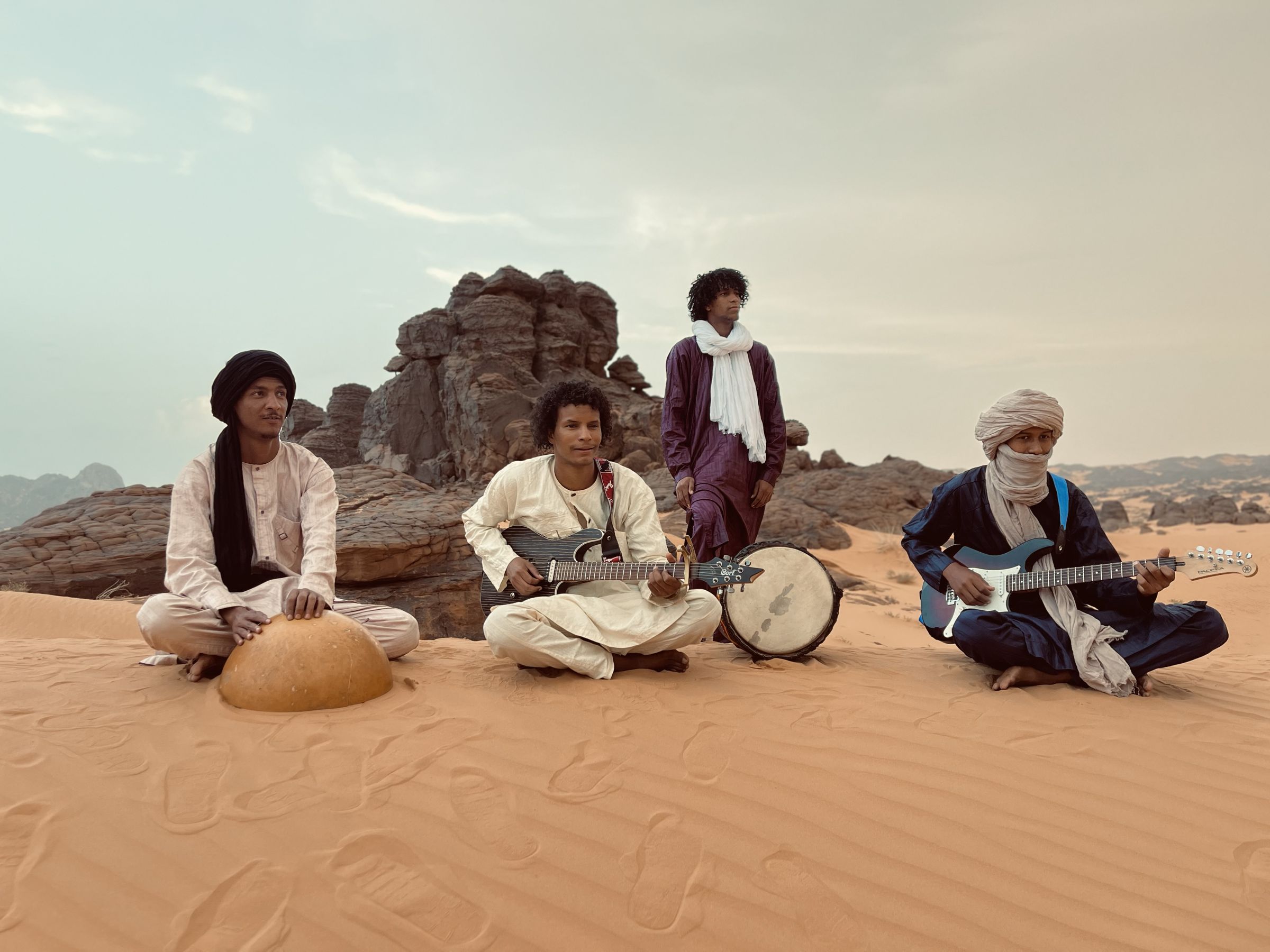 Los cuatro miembros de TAMODRE sentados en la arena frente a las rocas del Tassili, con guitarras eléctricas y calabaza